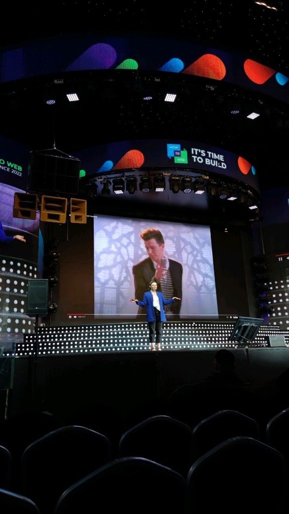 I'm shrugging on a stage in front of Rick Astley's music video "Never Gonna Give You Up." I'm wearing black wide legged cropped jeans, blue blazer and a white tank top.