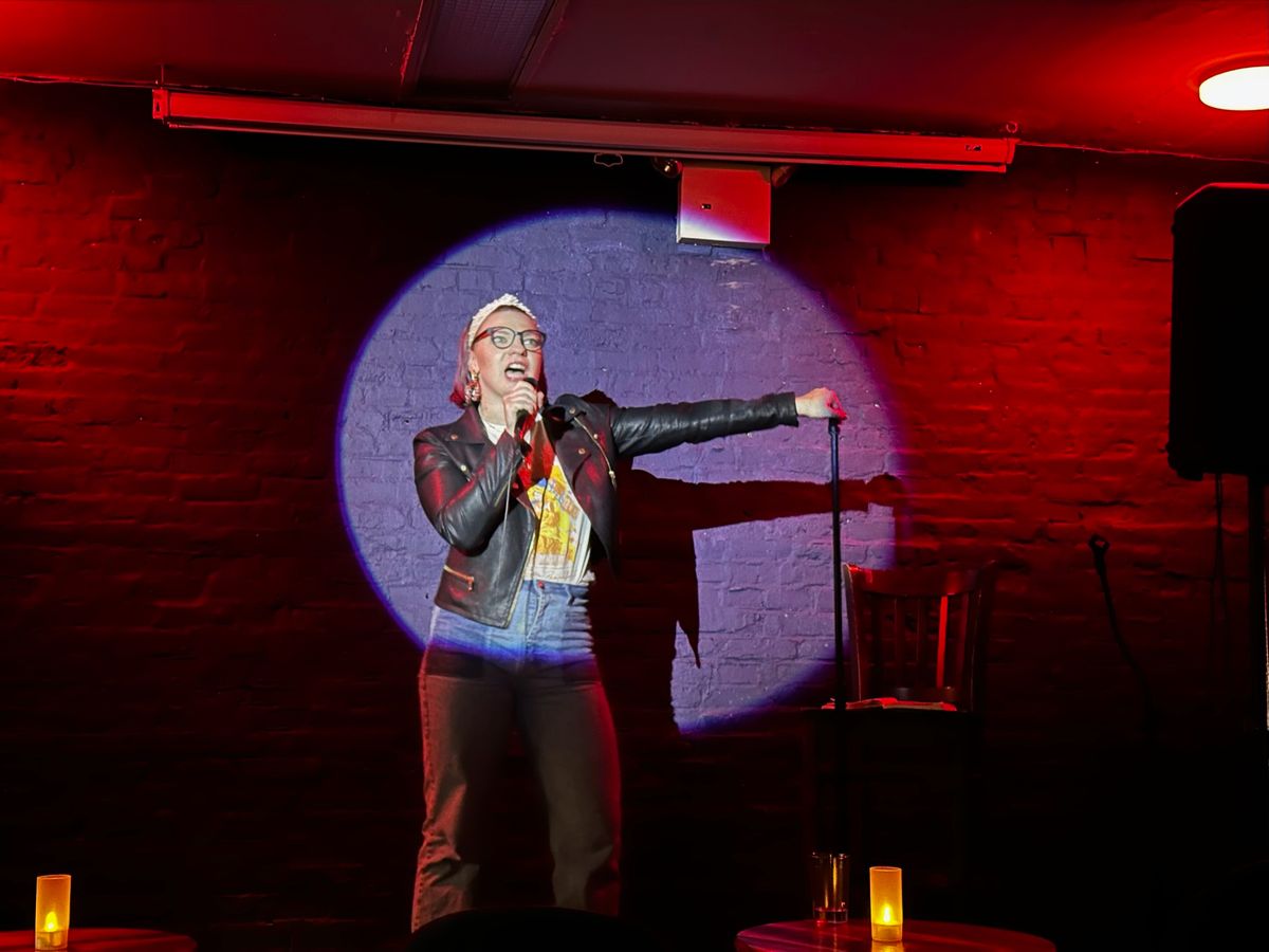A purple-haired woman in a headband and black leather jacket and high waisted jeans takes the stage and performs comedy